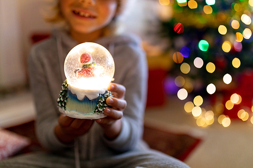 Front view mid section of a happy young Caucasian boy sitting on the floor holding a snow globe and smiling, in his sitting room at Christmas time