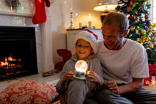 Front view of a happy middle aged Caucasian father sitting on the floor with his young son holding a snow globe and wearing a Santa hat, in their sitting room at Christmas time