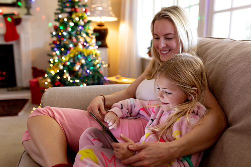 Side view of a happy young Caucasian mother sitting on the sofa with her young daughter using a tablet in their sitting room at Christmas time
