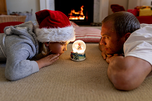 Side view of a middle aged Caucasian father lying on the floor with his young son looking at a snow globe together in their sitting room at Christmas time