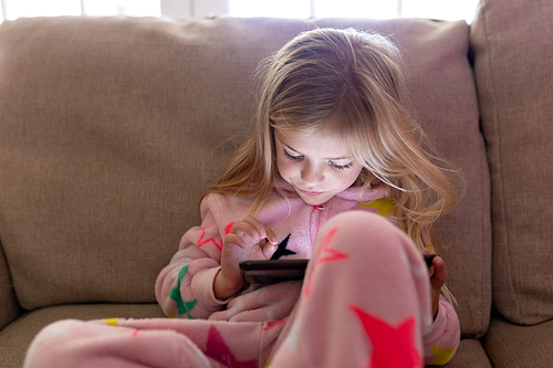 Front view close up of a happy young Caucasian girl sitting on a sofa wearing a onesie using a tablet in her sitting room at Christmas time