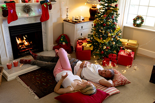High angle view of a Caucasian couple lying on the floor by the fireplace in their sitting room at Christmas time, looking at each other, holding hands and smiling, with a decorated Christmas tree in the background