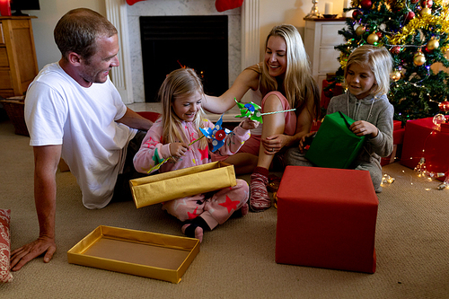 Front view of a Caucasian couple sitting on the floor with their young son and daughter in their sitting room at Christmas time opening presents and smiling by a decorated Christmas tree
