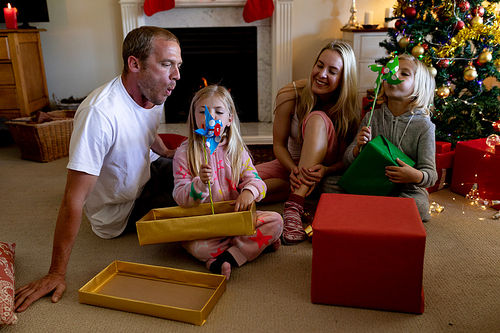 Front view of a Caucasian couple sitting on the floor with their young son and daughter in their sitting room at Christmas time, opening presents and playing with windmills