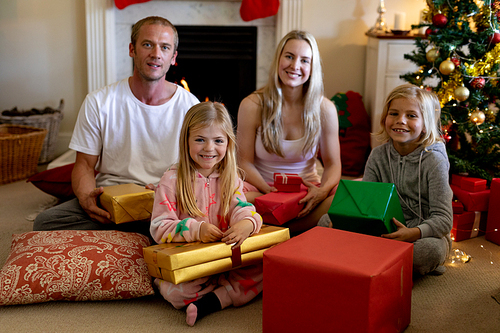 Portrait of a Caucasian couple sitting on the floor with their young son and daughter in their sitting room at Christmas time holding presents and smiling, a decorated Christmas tree in the background