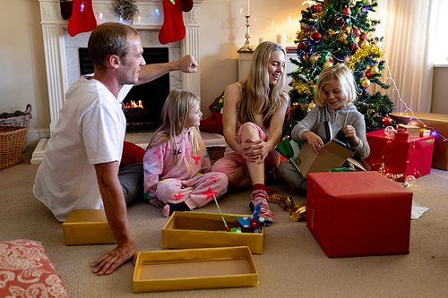 Front view of a Caucasian couple sitting on the floor with their young son and daughter in their sitting room at Christmas time, opening presents and smiling