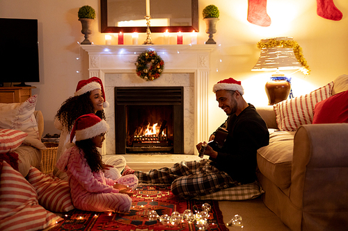 Side view of a mixed race couple sitting on the floor with their young daughter in their sitting room at Christmas, wearing santa hats, smiling, the man is playing guitar