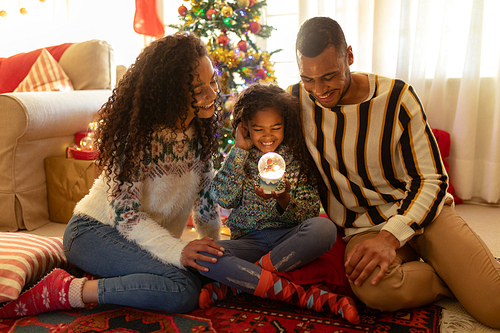 Front view of a mixed race couple with their young daughter in their sitting room at Christmas, sitting together on the floor smiling and looking at a snow globe held by the girl