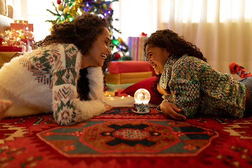 Side view of a mixed race woman with her young daughter in their sitting room at Christmas, lying on the floor facing each other with a snow globe in the middle and smiling