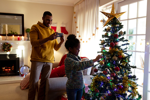 Front view of a mixed race man with his young daughter in their sitting room at Christmas, the man taking a photo of his daughter decorating a Christmas tree