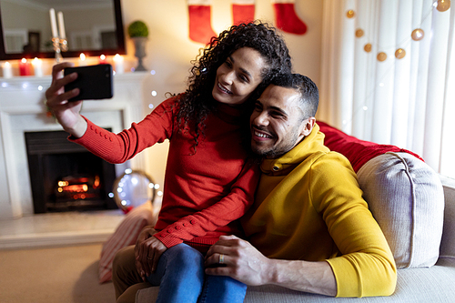 Front view of a mixed race couple sitting on a sofa in their sitting room at Christmas, taking a selfie