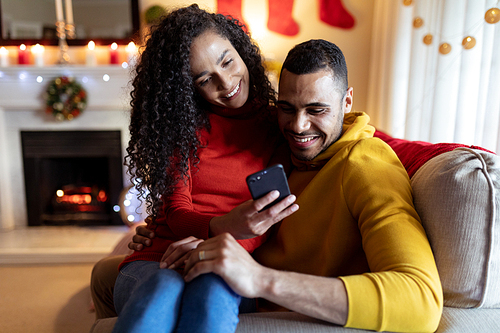 Front view of a mixed race couple sitting on a sofa in their sitting room at Christmas, using a smartphone