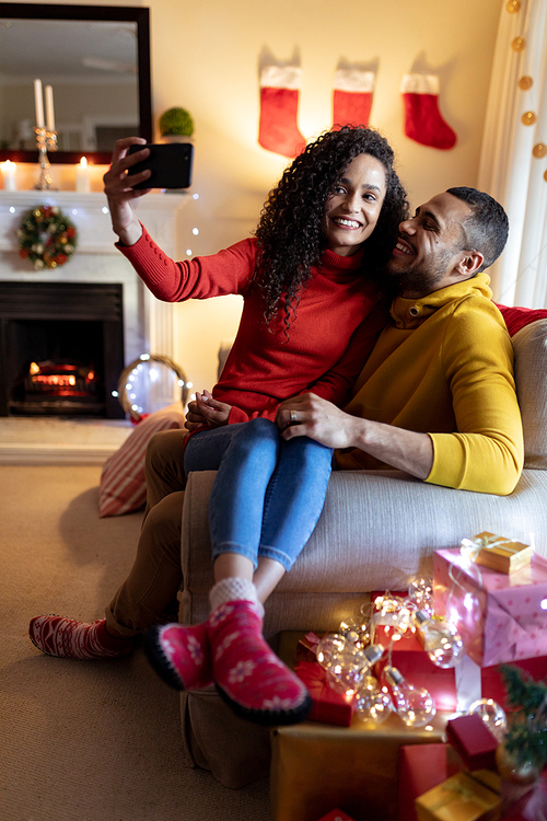 Front view of a mixed race couple sitting on a sofa in their sitting room at Christmas, taking a selfie