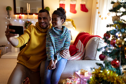 Front view of a mixed race man sitting on a sofa with his young daughter in their sitting room at Christmas, taking a selfie