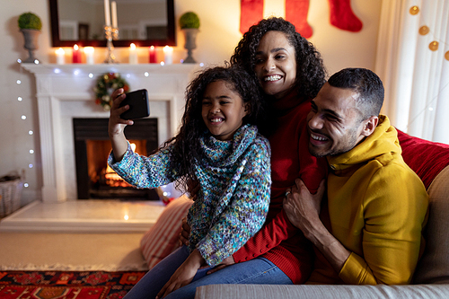 Front view of a mixed race couple with their young daughter in their sitting room at Christmas, sitting together on a sofa and taking a selfie