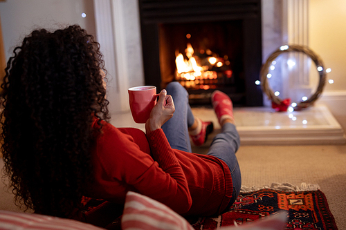 Rear view of a mixed race woman in her sitting room at Christmas, lying by a fireplace and holding a mug