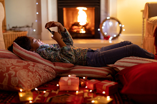 Side view of a young mixed race girl in her sitting room at Christmas, lying on cushions on the floor and using a smartphone