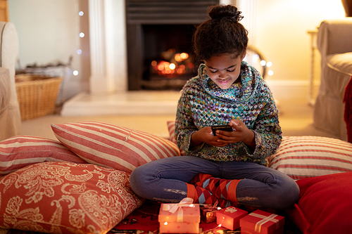 Front view of a young mixed race girl in her sitting room at Christmas, lying on cushions on the floor and using a smartphone