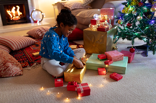 Side view of a young mixed race girl sitting on the floor in her sitting room at Christmas, smiling and opening a present