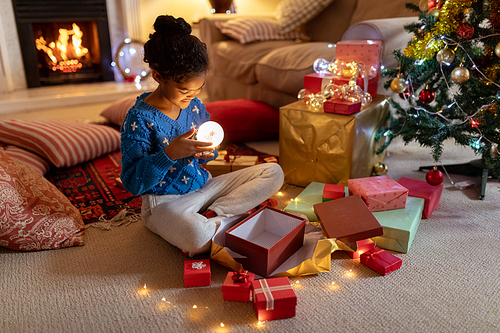Side view of a young mixed race girl sitting on the floor in her sitting room at Christmas, smiling and holding a snow globe