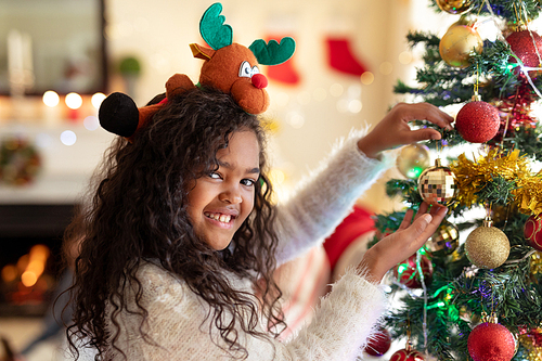 Portrait of a young mixed race girl in her sitting room at Christmas, wearing a festive hat holding a bauble on a Christmas tree smiling