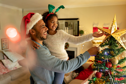 Side view of a mixed race man with his young daughter in their sitting room at Christmas, wearing festive hats, the man holding his daughter and touching a Christmas tree