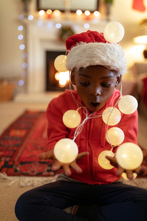 Front view of a young mixed race boy in his sitting room at Christmas, wearing a santa hat and holding fairy lights