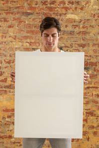 Front view close up of a young Caucasian man holding a blank white canvas in his hands and looking down at it, standing in front of a brick wall