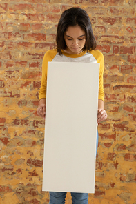 Front view close up of a young Caucasian woman holding a blank white canvas in her hands and flooking down at it, standing in front of a brick wall