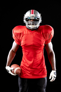 Portrait of an African American male American football player wearing a team uniform, pads and a helmet holding the ball in one hand. Vertical shot