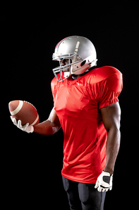 Side view close up of an African American male American football player wearing a team uniform, pads and a helmet, holding a football in one hand and looking down at it. Vertical shot