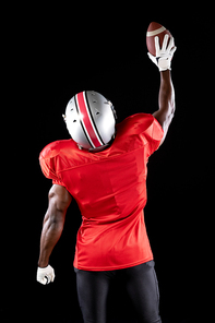 Rear view of an African American male American football player wearing a team uniform, pads, gloves and a helmet, with one arm raised holding a football. Vertical shot