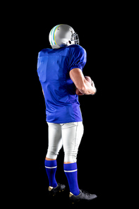 Rear view of a Caucasian male American football player wearing a team uniform, pads and a helmet, holding a football under his arm. Vertical shot