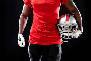 Front view mid section of an African American male American football player wearing a team uniform, pads and gloves, holding a helmet under his arm