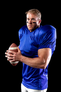 Side view of a Caucasian male American football player wearing a team uniform, pads and eye black under his eyes, holding a football in his hands and turning to camera. Vertical shot