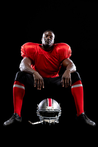 Portrait of an African American male American football player wearing a team uniform with his helmet on the ground in front of him, sitting looking to camera. Vertical shot