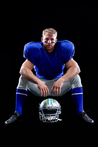 Portrait of a Caucasan male American football player wearing a team uniform with his helmet on the ground in front of him, sitting looking to camera. Vertical shot