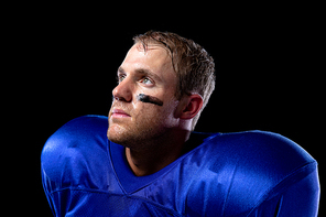 Side view close up of a Caucasian male American football player wearing a team uniform with shoulder pads and lines of eye black under his eyes, looking up