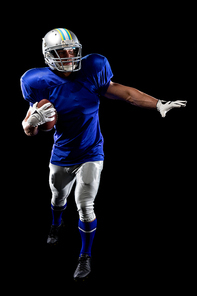 Front view of a Caucasian male American football player wearing a team uniform, pads and a helmet holding a football under his arm with the other arm outstretched. Vertical shot