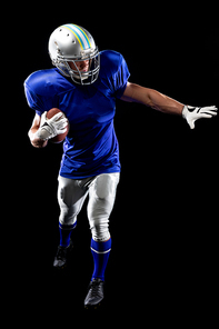 Front view of a Caucasian male American football player wearing a team uniform, pads and a helmet holding a football under his arm with the other arm outstretched. Vertical shot