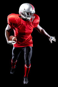 Front view of an African American male American football player wearing a team uniform, pads and a helmet, stepping forward with head turned away and a football in his hand and his other arm outstretched. Vertical shot