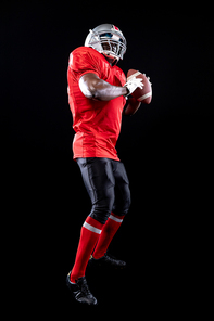 Side view close up of an African American male American football player wearing a team uniform, pads and a helmet, holding a football in both hands