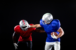 Front view of a Caucasian male American football player wearing team colours, pads and a helmet holding the football and pushing away an African American player on the opposing team