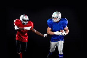 Front view of a Caucasian male American football player wearing team colours, pads and a helmet running holding a football pursued by an African American player on the opposing team