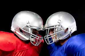 Side view close up of a Caucasian and an African American male American football player wearing helmets and their opposing team colours, standing opposite staring at each other, their helmets touching