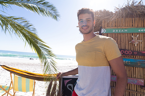Caucasian man enjoying time at the beach, standing by a surf shop, looking at the camera and smiling