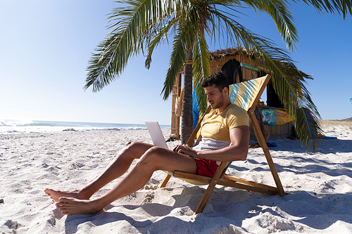 Caucasian man enjoying time at the beach, sitting on a deck chair, using a laptop