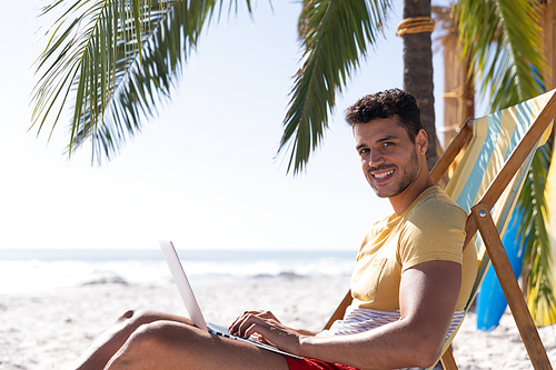 Caucasian man enjoying time at the beach, sitting on a deck chair, using a laptop