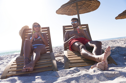 Caucasian couple enjoying time at the beach on a sunny day, sitting on deck chairs and drinking drinks with sea in the background