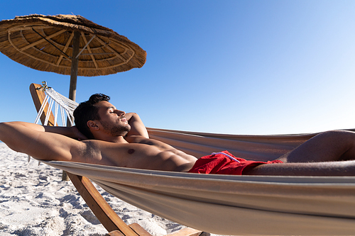 Caucasian man enjoying time at the beach on a sunny day, lying on a hammock and sunbathing with sea in the background